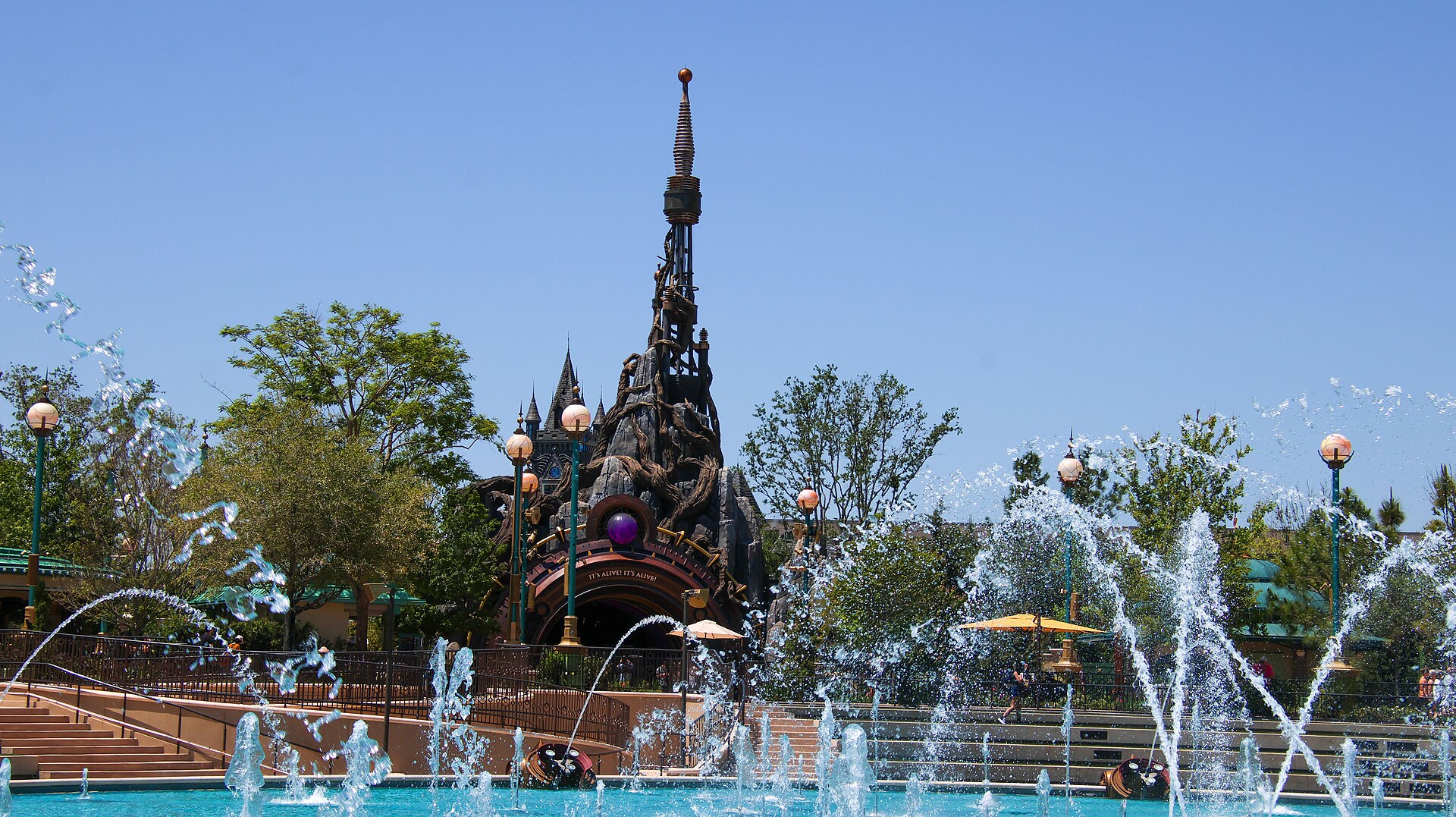 The gnarled rock and tree portal to Dark Universe seen across the fountains of Celestial Park, with Frankenstein Manor's spire rising behind it
