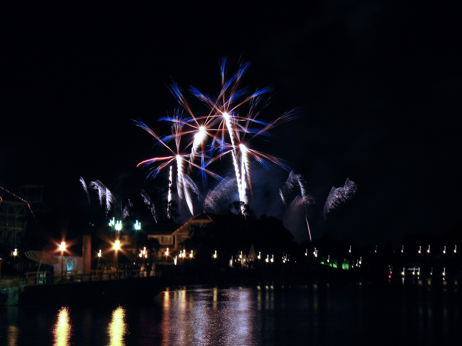 Fireworks rising above trees and buildings beyond the water, seen from Disney's BoardWalk at night