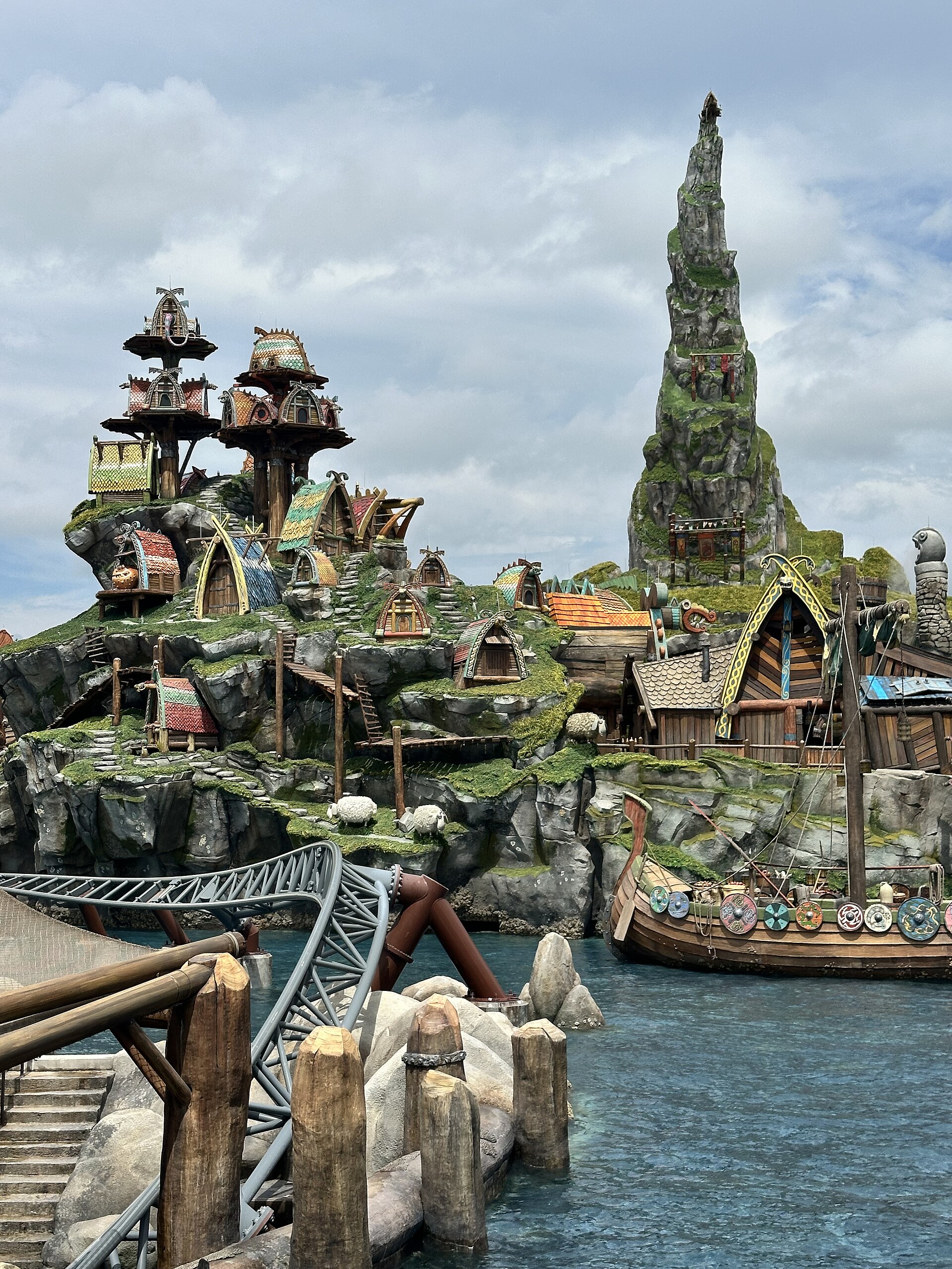 The Viking village of Berk at Epic Universe, with colourful longhouses on a green hillside, a sea stack and a longship moored on the water