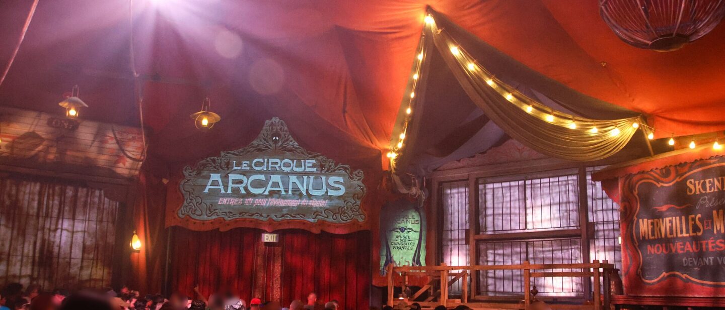 The red pre-show tent for Le Cirque Arcanus in the Ministry of Magic, with a painted sign, strings of lights and an audience waiting