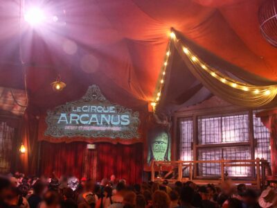 The red pre-show tent for Le Cirque Arcanus in the Ministry of Magic, with a painted sign, strings of lights and an audience waiting
