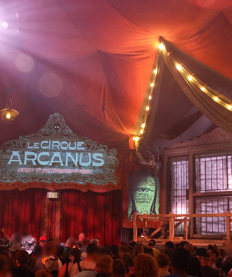 The red pre-show tent for Le Cirque Arcanus in the Ministry of Magic, with a painted sign, strings of lights and an audience waiting