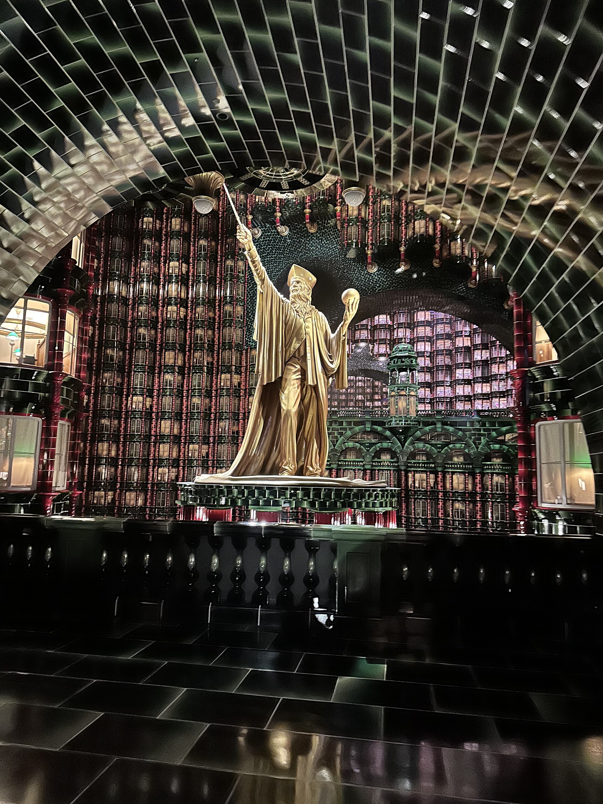 A towering golden wizard statue in the Ministry of Magic atrium, framed by green-tiled arches and walls of windows