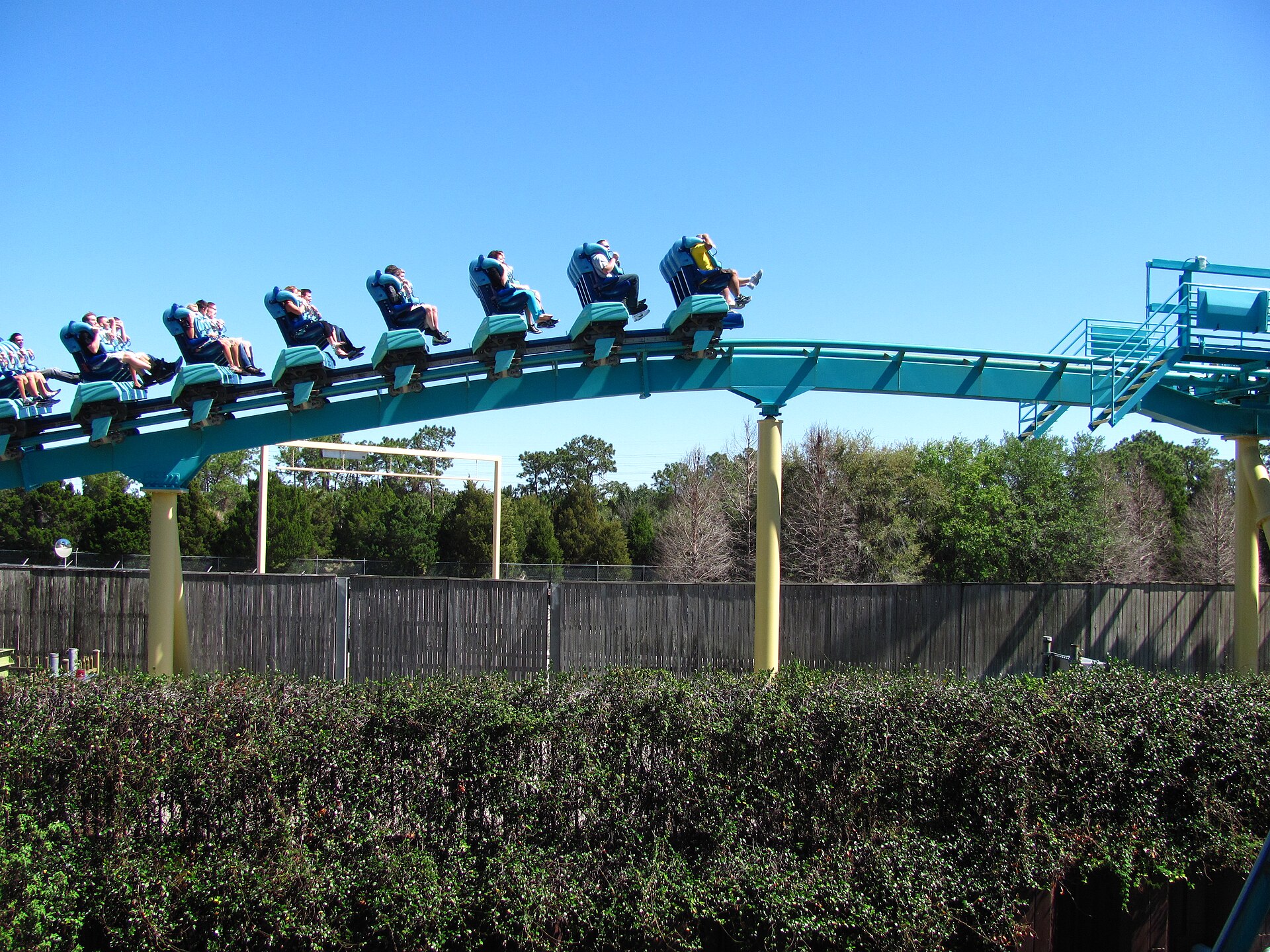 A full train of riders on the teal Kraken floorless coaster track against a blue sky at SeaWorld Orlando