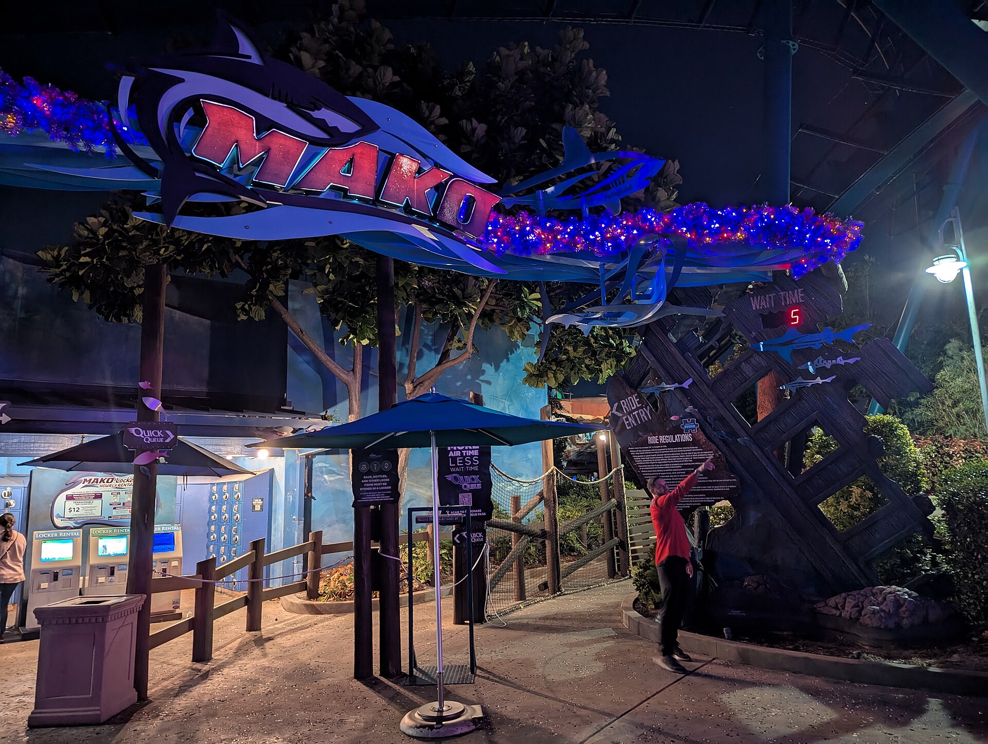 Mako roller coaster entrance sign at night, lit in blue and red and trimmed with festive tinsel garland