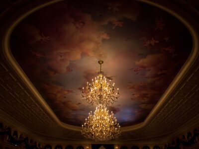 Two tiers of lit chandeliers hanging from a painted ceiling of clouds and cherubs in the ballroom at Be Our Guest Restaurant