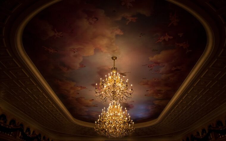 Two tiers of lit chandeliers hanging from a painted ceiling of clouds and cherubs in the ballroom at Be Our Guest Restaurant