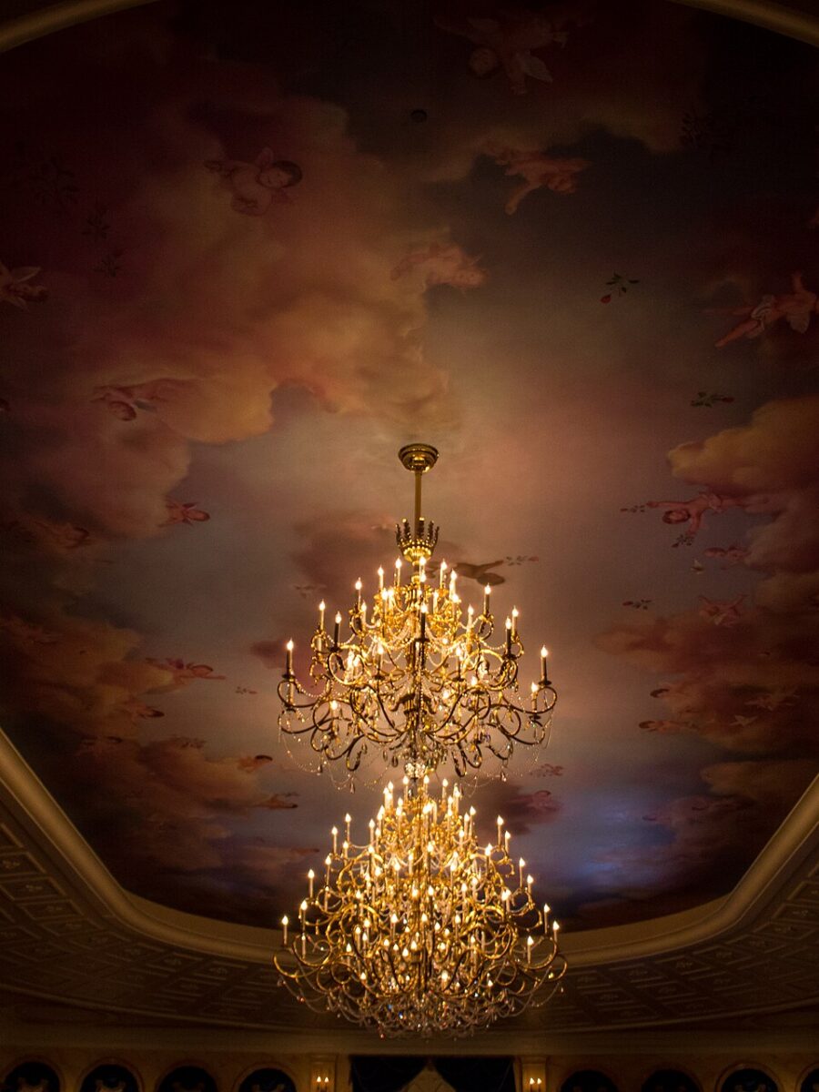 Two tiers of lit chandeliers hanging from a painted ceiling of clouds and cherubs in the ballroom at Be Our Guest Restaurant
