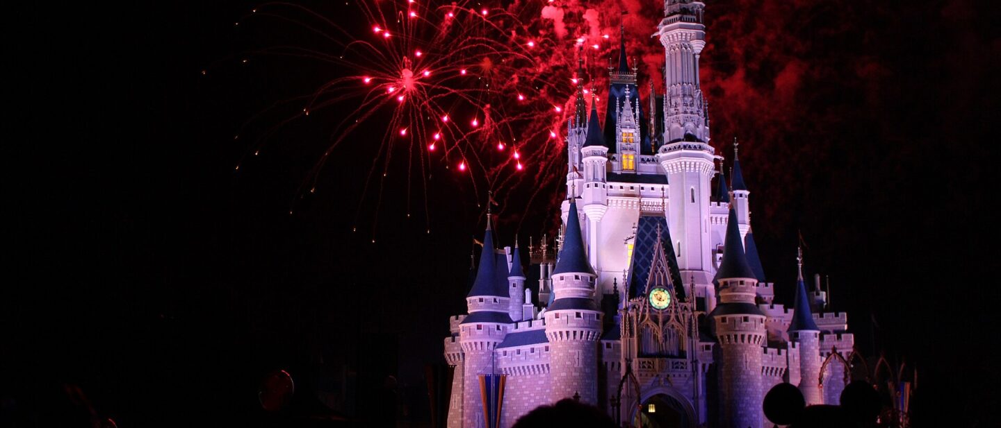Cinderella Castle lit white and blue at night with red fireworks bursting above it and the silhouettes of a crowd in the foreground