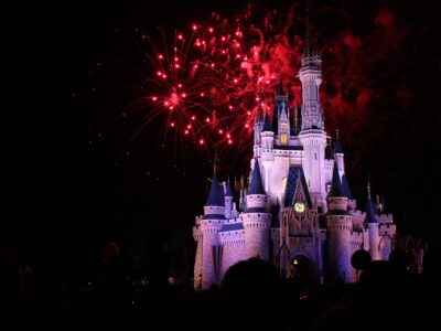 Cinderella Castle lit white and blue at night with red fireworks bursting above it and the silhouettes of a crowd in the foreground