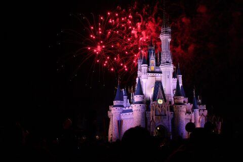 Cinderella Castle lit white and blue at night with red fireworks bursting above it and the silhouettes of a crowd in the foreground