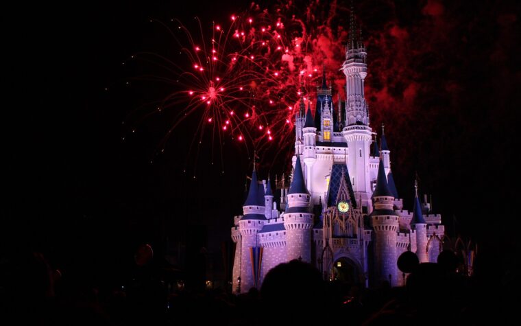 Cinderella Castle lit white and blue at night with red fireworks bursting above it and the silhouettes of a crowd in the foreground