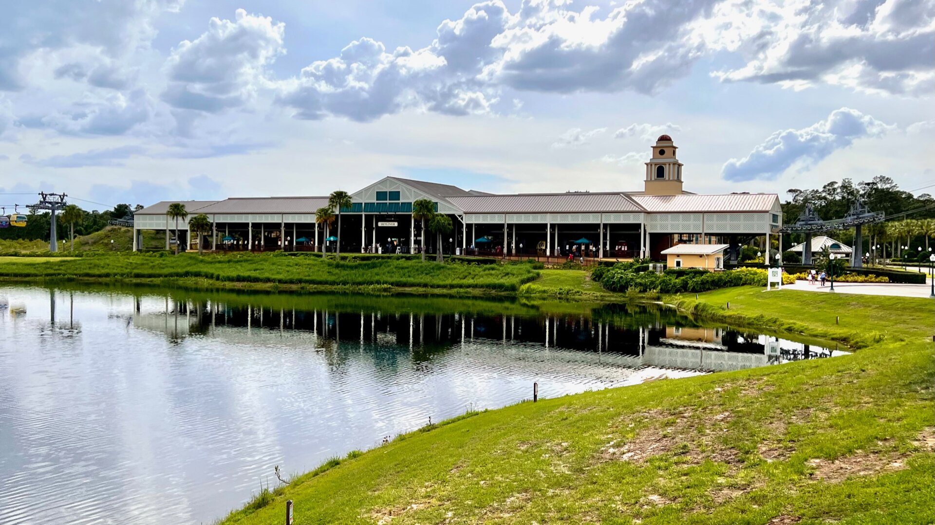 The Disney Skyliner station at Caribbean Beach Resort, a long open-sided terminal with a clock tower, reflected in a lake under a cloudy sky