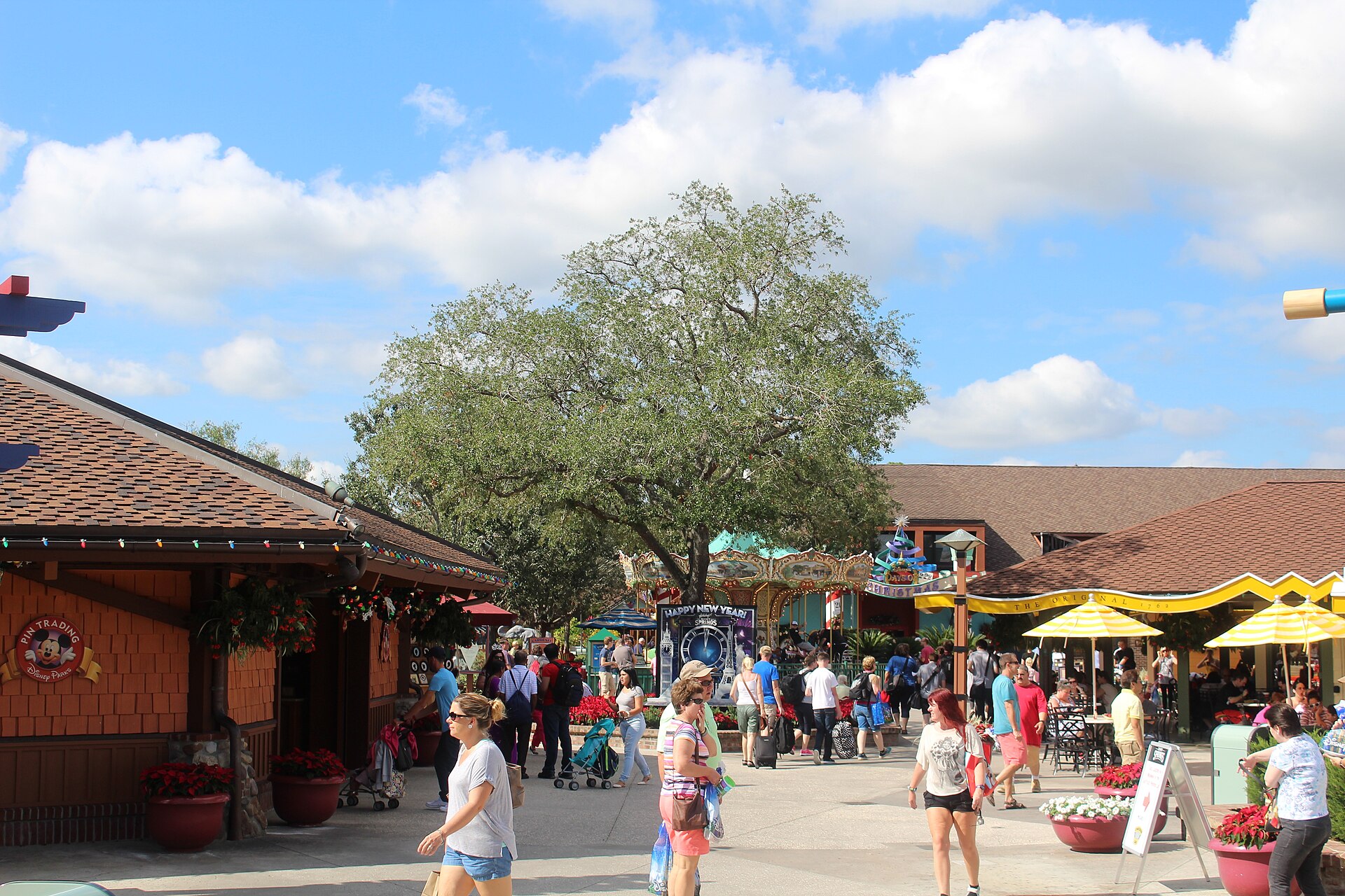 Shoppers walking past wooden-roofed shops and restaurants under a large oak tree at Disney Springs on a sunny day
