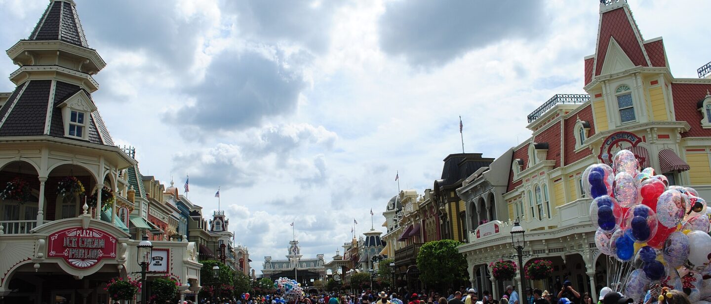 Crowds walking down Main Street, U.S.A. at Magic Kingdom towards Main Street station under a cloudy sky, with a balloon seller on the right