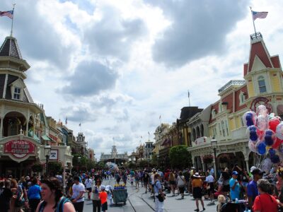 Crowds walking down Main Street, U.S.A. at Magic Kingdom towards Main Street station under a cloudy sky, with a balloon seller on the right
