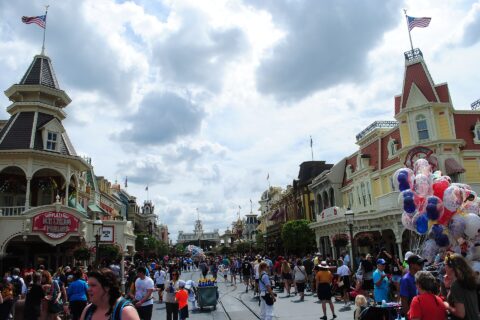 Crowds walking down Main Street, U.S.A. at Magic Kingdom towards Main Street station under a cloudy sky, with a balloon seller on the right