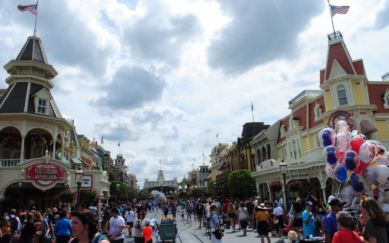 Crowds walking down Main Street, U.S.A. at Magic Kingdom towards Main Street station under a cloudy sky, with a balloon seller on the right