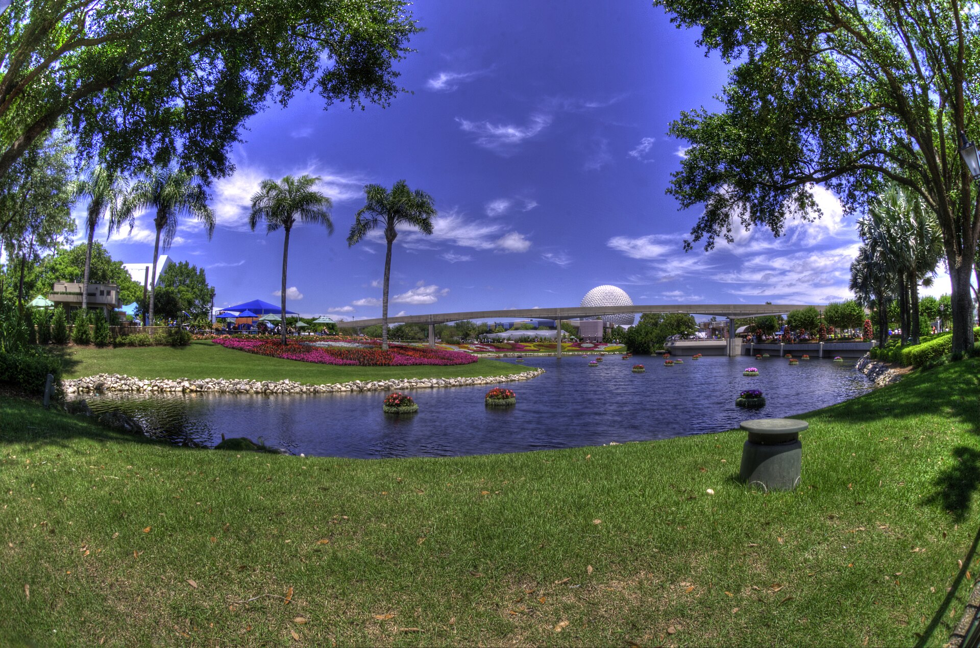 Flower beds and palm trees around the lagoon at EPCOT, with Spaceship Earth and the monorail track in the distance under a blue sky