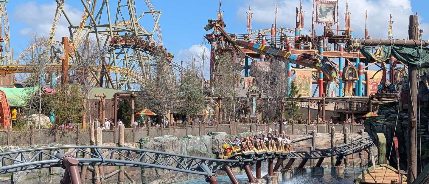 A Hiccup's Wing Gliders train full of riders with raised arms skims over the lagoon in Isle of Berk, with the coaster's taller track and the striped arms of Dragon Racer's Rally behind
