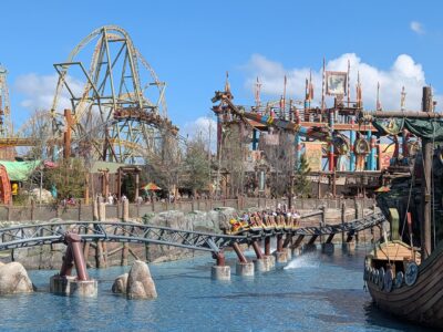 A Hiccup's Wing Gliders train full of riders with raised arms skims over the lagoon in Isle of Berk, with the coaster's taller track and the striped arms of Dragon Racer's Rally behind