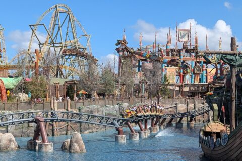A Hiccup's Wing Gliders train full of riders with raised arms skims over the lagoon in Isle of Berk, with the coaster's taller track and the striped arms of Dragon Racer's Rally behind