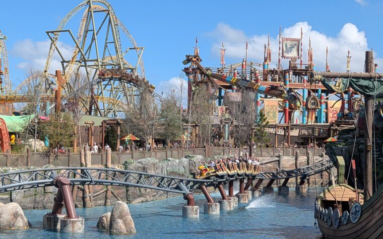 A Hiccup's Wing Gliders train full of riders with raised arms skims over the lagoon in Isle of Berk, with the coaster's taller track and the striped arms of Dragon Racer's Rally behind