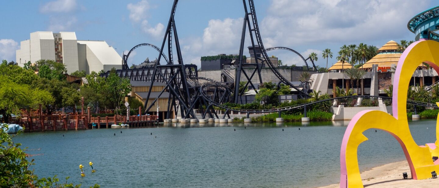 VelociCoaster's black top hat rising over the Islands of Adventure lagoon, with Jurassic Park's thatched buildings to the right and Seuss Landing's colorful arches in the foreground