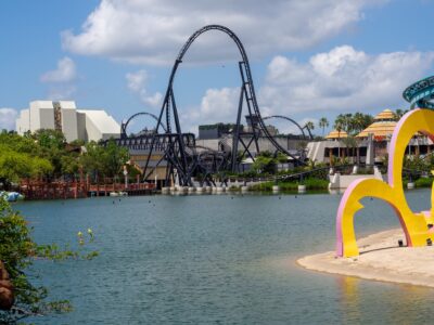 VelociCoaster's black top hat rising over the Islands of Adventure lagoon, with Jurassic Park's thatched buildings to the right and Seuss Landing's colorful arches in the foreground