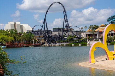 VelociCoaster's black top hat rising over the Islands of Adventure lagoon, with Jurassic Park's thatched buildings to the right and Seuss Landing's colorful arches in the foreground