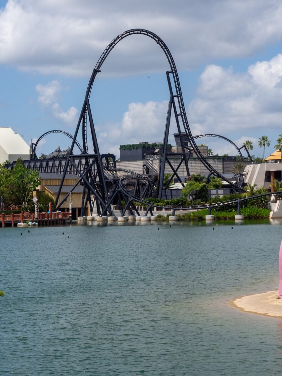 VelociCoaster's black top hat rising over the Islands of Adventure lagoon, with Jurassic Park's thatched buildings to the right and Seuss Landing's colorful arches in the foreground