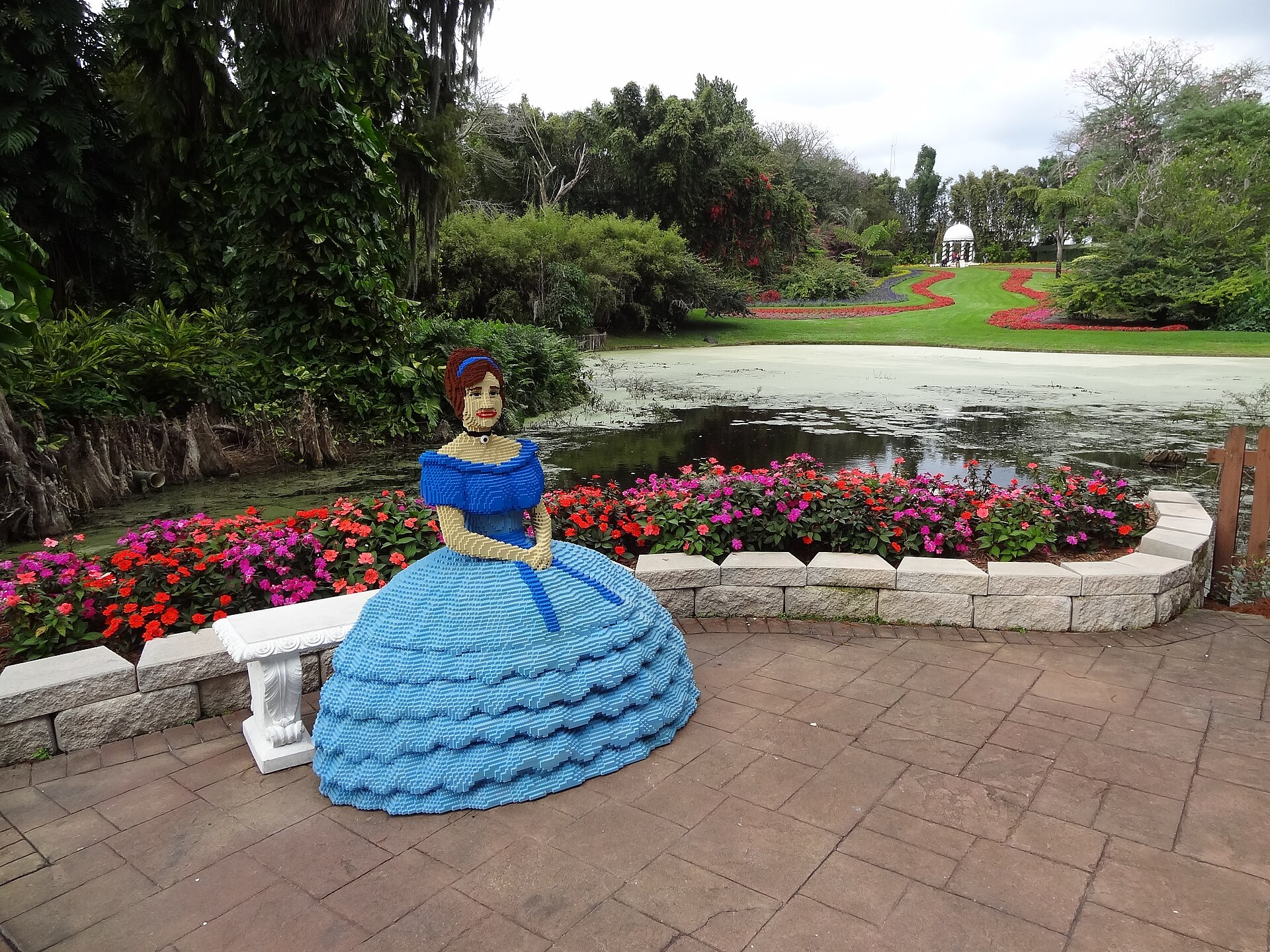 A life-size LEGO figure of a Southern belle in a blue gown seated beside a flower-lined pond in the gardens at LEGOLAND Florida