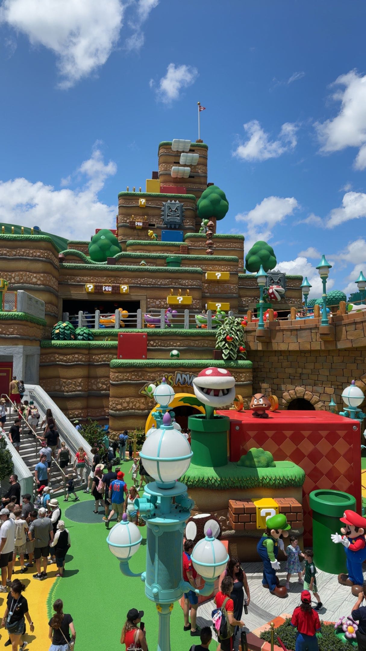 The Yoshi's Adventure entrance in Super Nintendo World at Epic Universe, with a Piranha Plant and a Goomba on the facade, Yoshi ride vehicles on the mountain above and Mario and Luigi greeting guests in the foreground