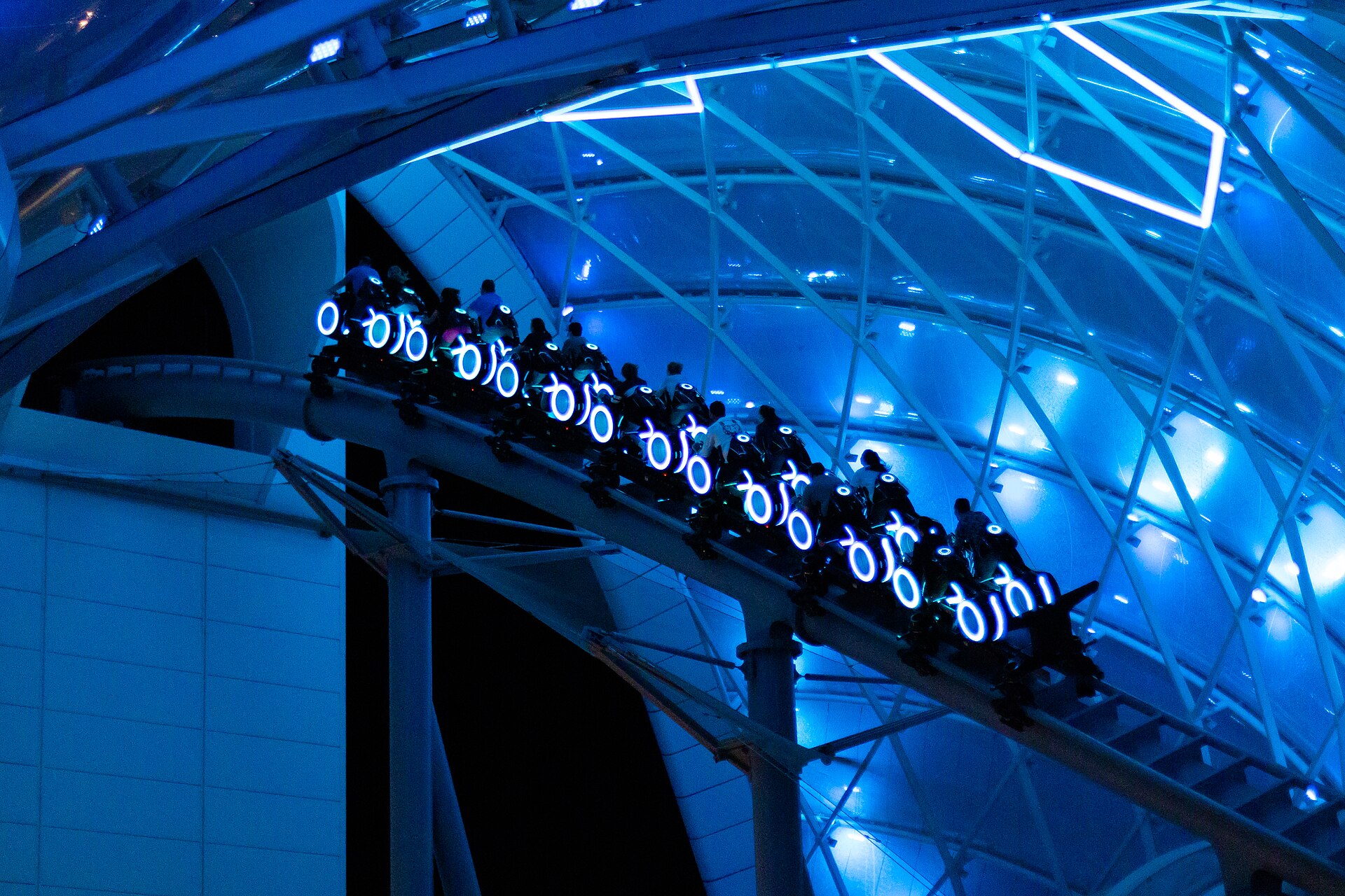 TRON Lightcycle Run ride vehicles lit in blue rings speeding under the illuminated canopy at night