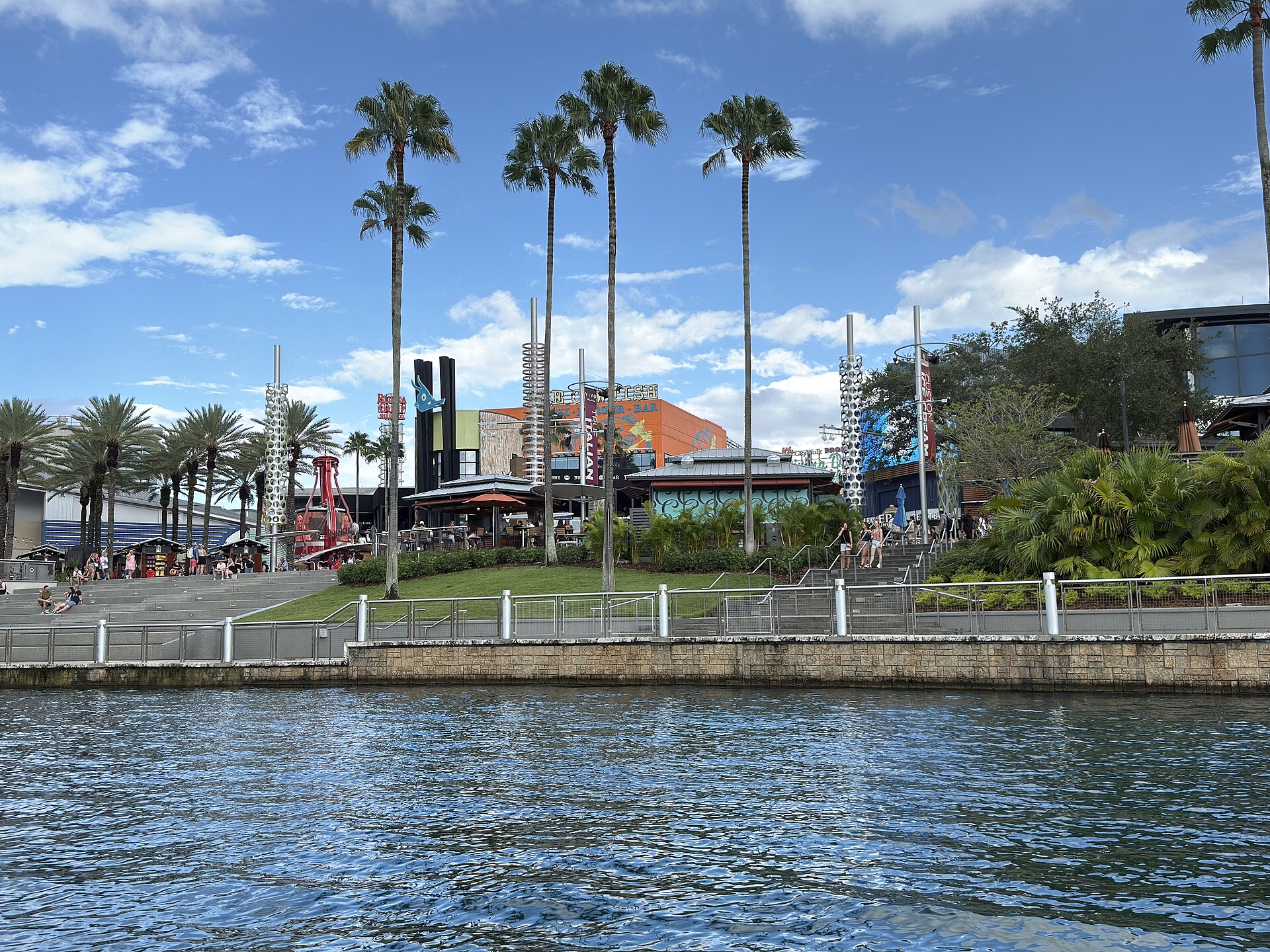 Universal CityWalk seen across the lagoon on a sunny day, with tall palm trees, restaurant buildings and a landscaped promenade